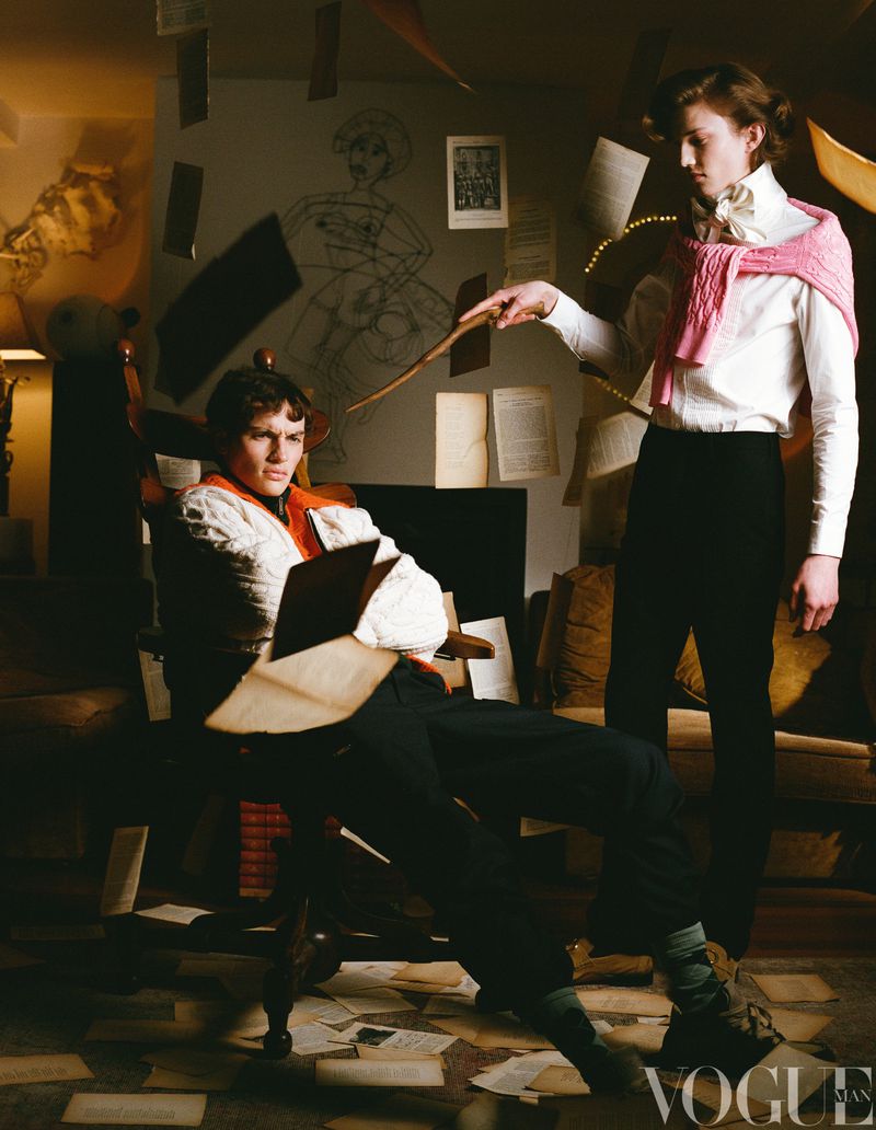 Two adult males in vintage fashion, one seated with papers and books, the other standing with a wand in an indoor setting filled with papers.