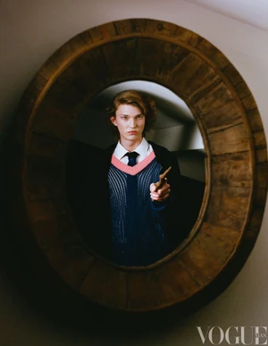 Young model holding a small wand, reflected inside a large round wooden-framed mirror wearing a sweater vest and shirt