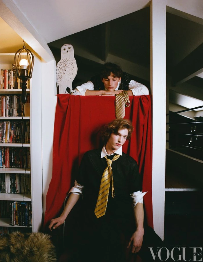 Two male models in school shirts and ties posing in an interior space with a red drapery and an owl figure.