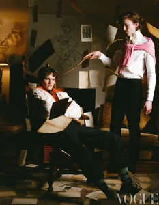 Two male models in a room with floating papers, one standing holding a wand and one seated in a chair.