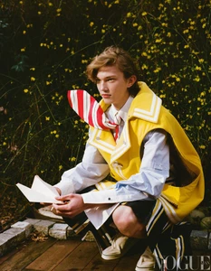 Young male model crouching outdoors holding a paper airplane wearing a yellow vest and striped tie
