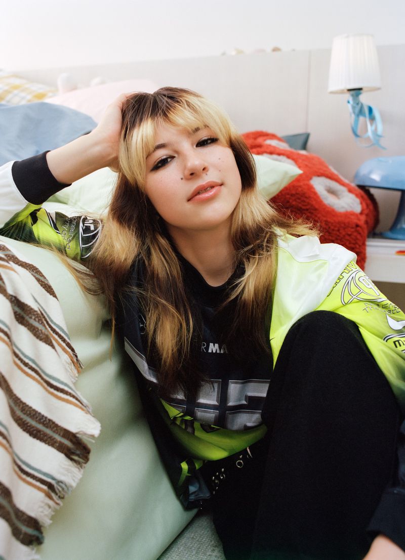 Portrait of a young woman with blonde bangs and a graphic t-shirt, posing indoors in a bedroom setting with decorative pillows.