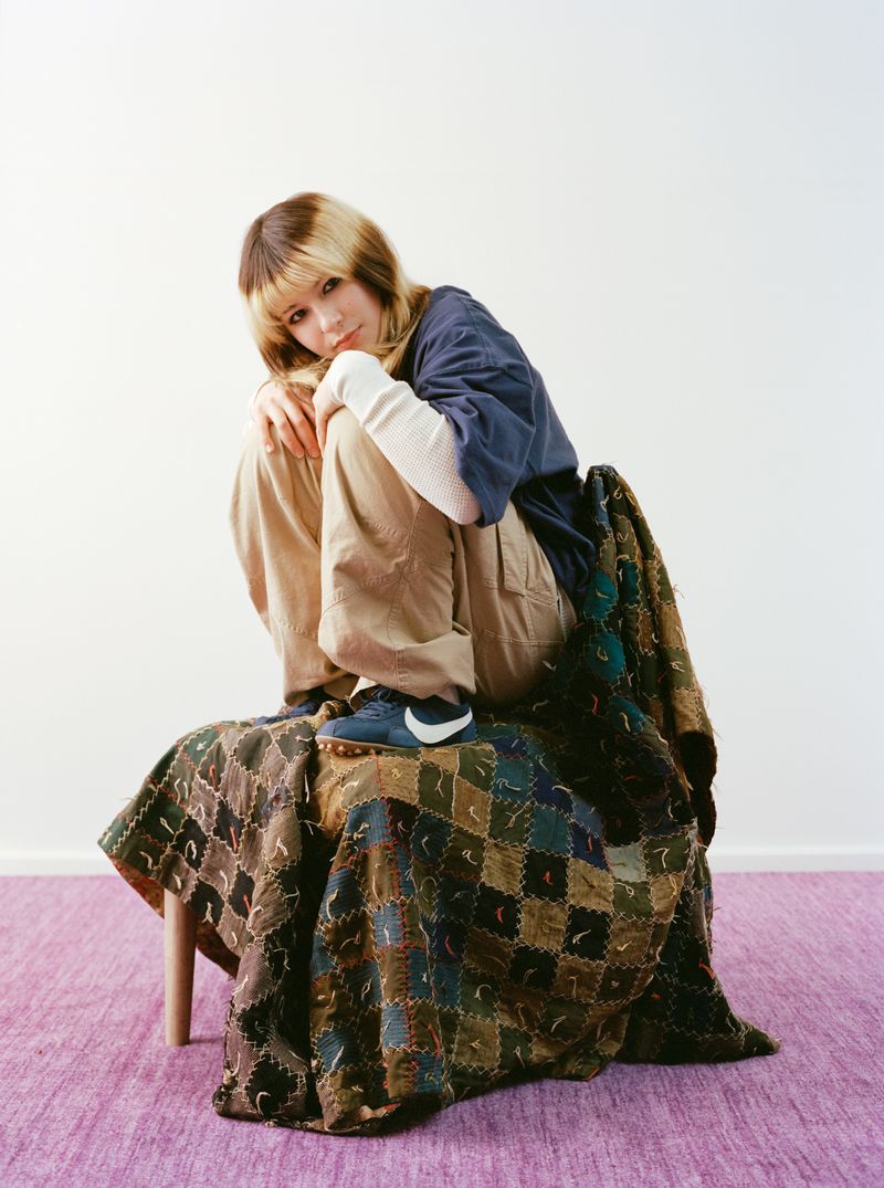 A woman with brown and blonde hair is sitting on a blanket-covered chair in a studio with a white wall and purple rug.