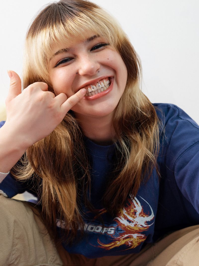 Smiling woman in casual clothing makes a 'call me' gesture, showcasing dental braces. Indoor portrait with a white background.