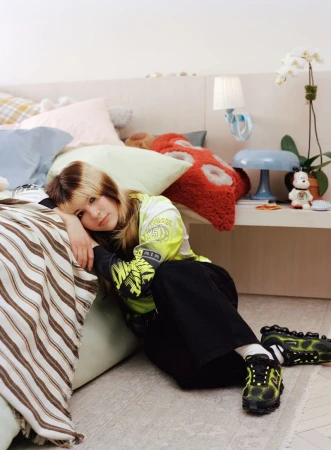 A young woman with blond hair and black pants poses with a yellow shirt on the floor beside a striped bed and lamp in an indoor setting.
