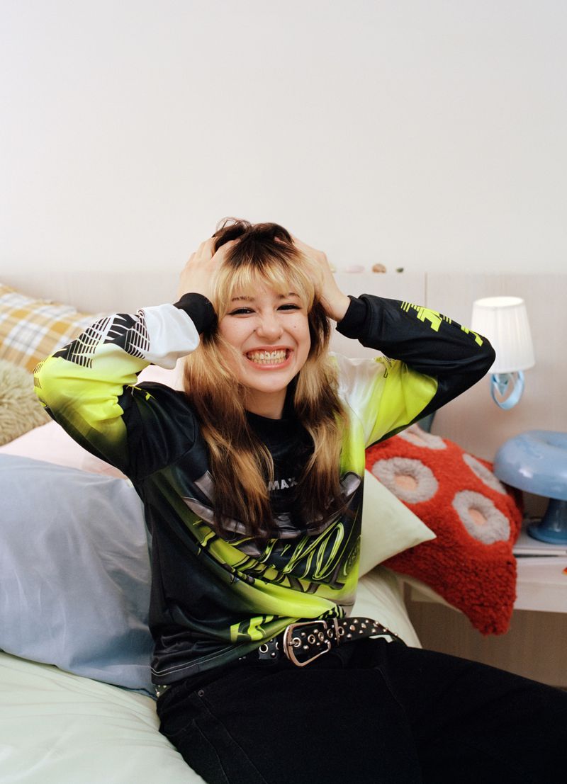 Indoor studio shot of a young adult woman with hands in her hair, wearing a graphic print long-sleeve shirt, and smiling at the camera.