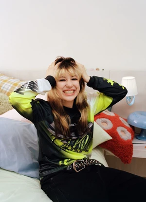 Alysa Liu sitting on a bed smiling with hands on head, wearing a graphic long-sleeve top and black studded belt