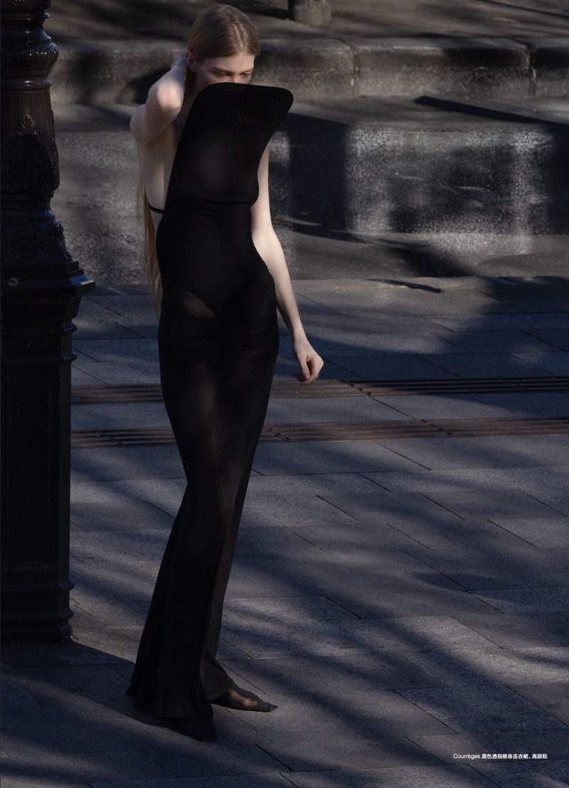 Fashion shot of an adult woman wearing a long black dress standing outside on a stone surface in dappled sunlight.