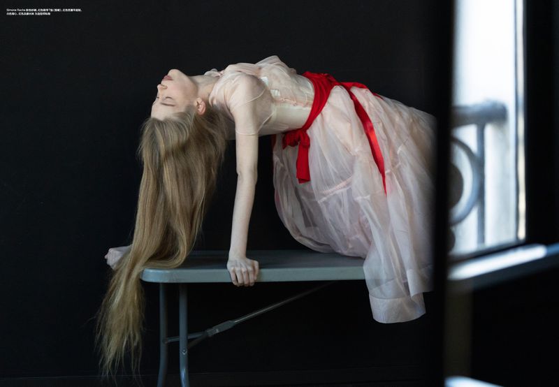 Fashion shot of a young adult female model in a light pink dress with a red sash, leaning on a table with long hair, against a dark background.