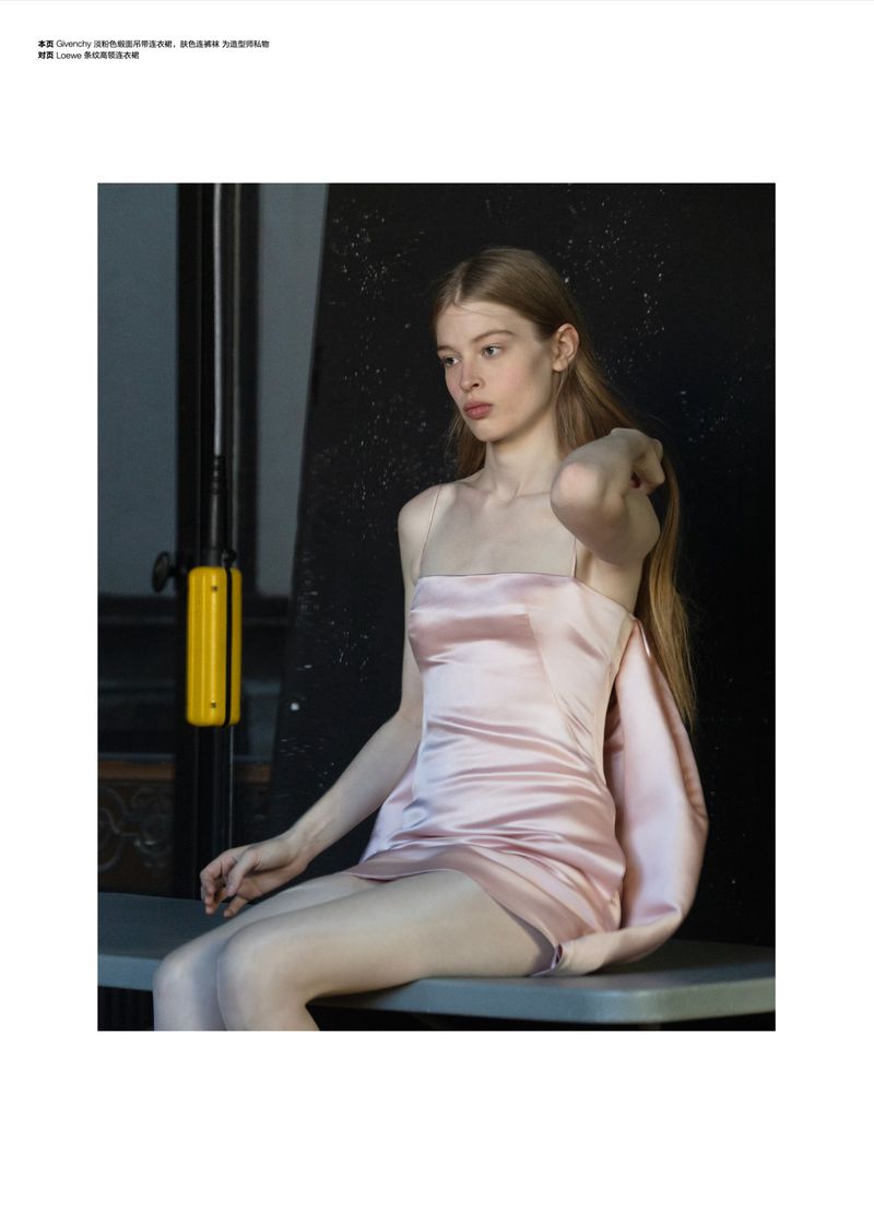Young woman in light pink dress seated on a gray surface, glancing slightly down, with dark and muted lighting, fashion portrait.