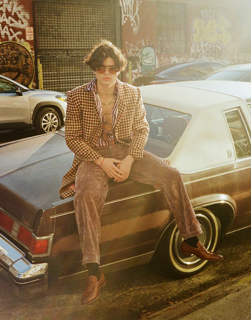 Adult male model wearing sunglasses and a checkered blazer poses on a classic car in an urban street setting with natural light.