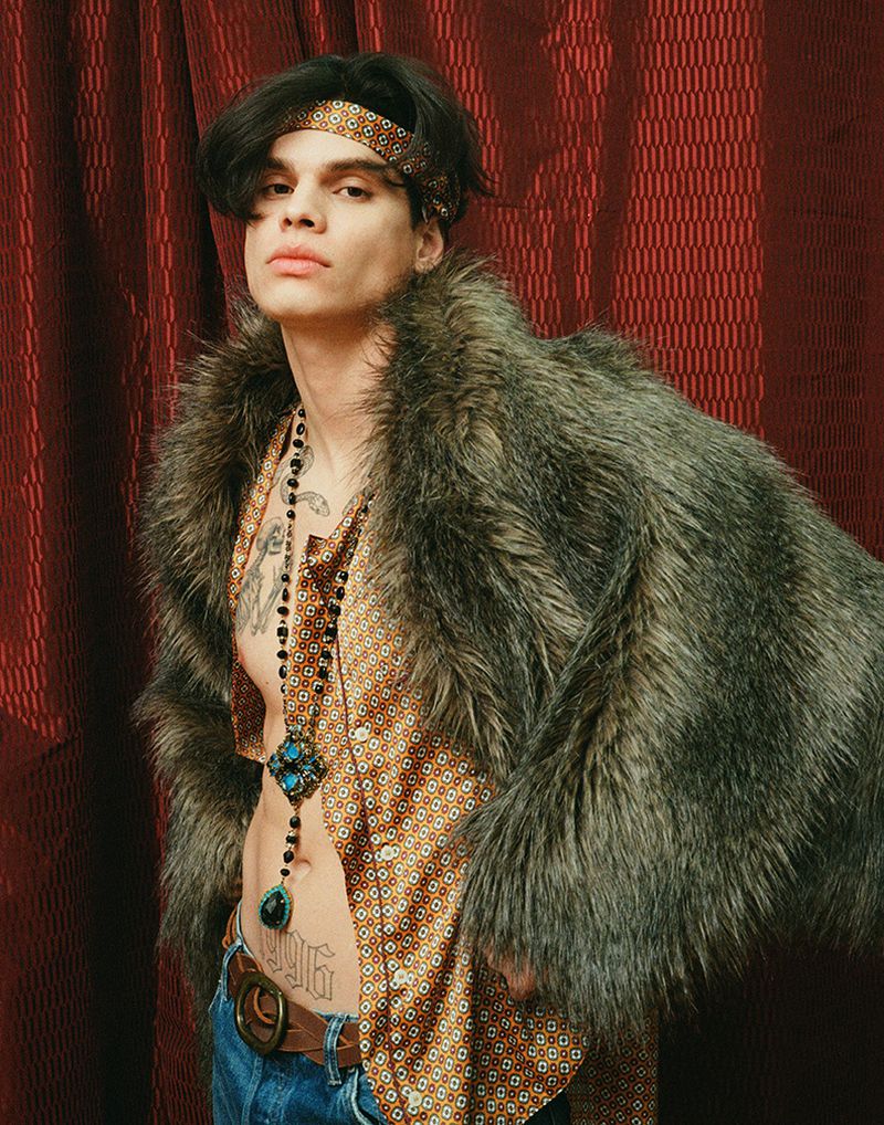 Young man with dark hair wears a geometric print shirt, faux fur coat, headscarf, and long beaded pendant necklace against a red curtain.