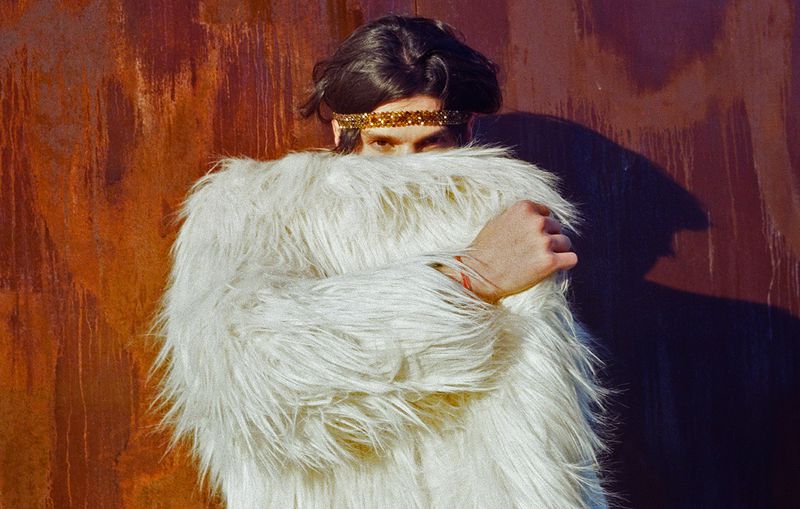 A person with dark hair wears a sparkly gold headband and a long-haired white faux fur coat, clutching it close against a rusty metal wall.