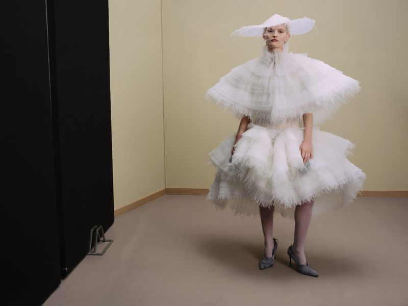 Full-length shot of a model in a surreal, oversized white couture outfit with a hat, set against a minimalist backdrop.