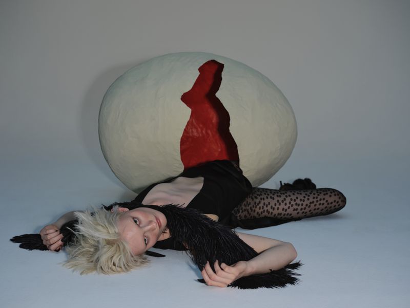 A woman lies next to a large egg prop, wearing a black outfit with a feathered collar and patterned tights, in a photo studio setting.