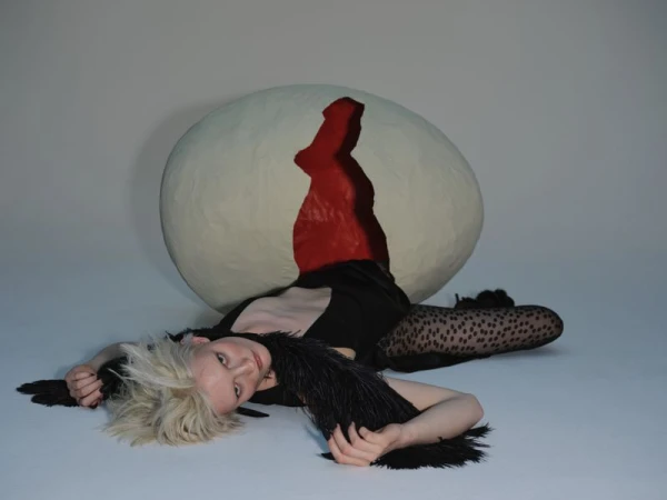 A woman lies next to a large egg prop, wearing a black outfit with a feathered collar and patterned tights, in a photo studio setting.