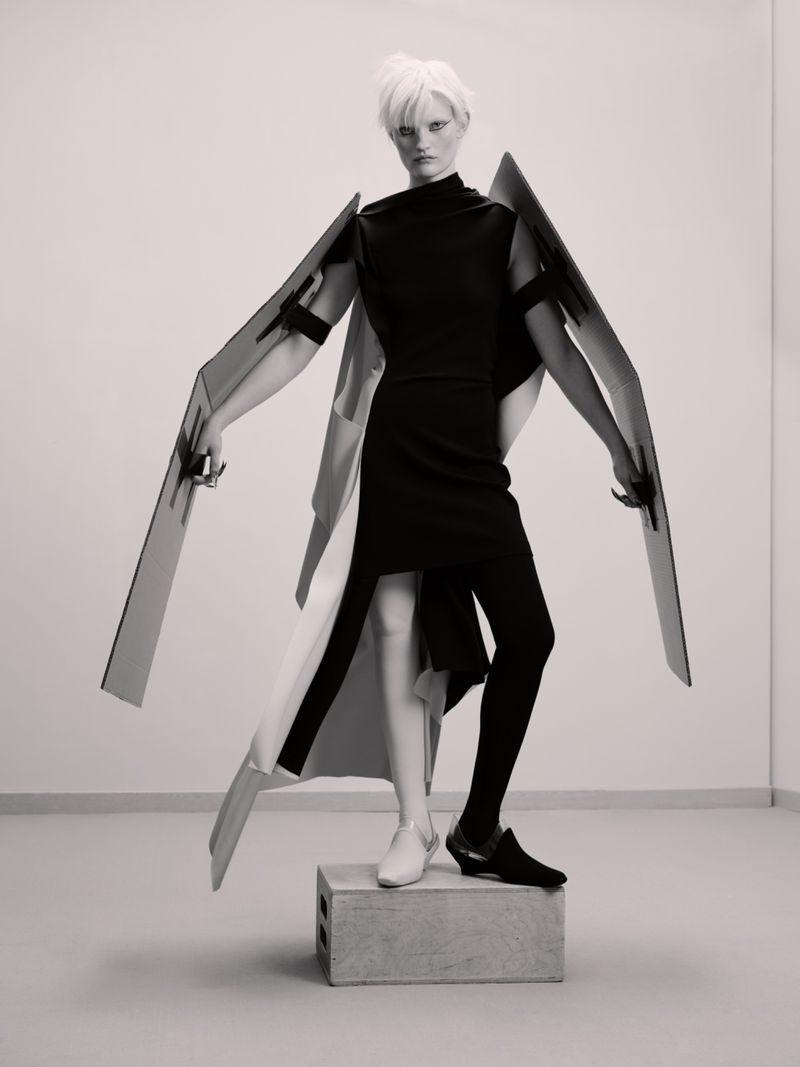 Fashion photo of a person with short white hair wearing a black dress and black leggings, standing on a concrete block in a studio.