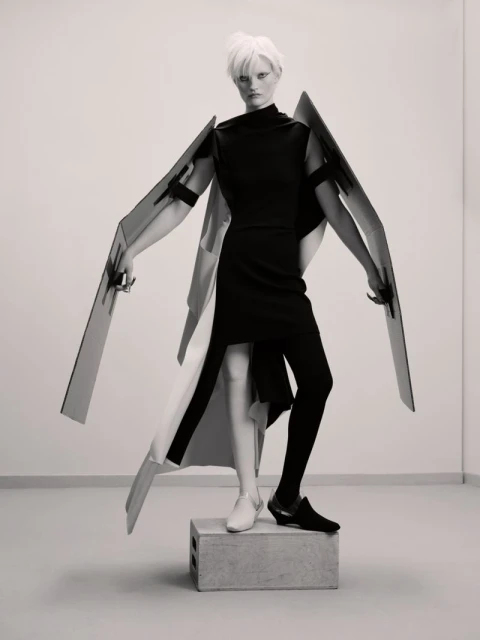 Fashion photo of a person with short white hair wearing a black dress and black leggings, standing on a concrete block in a studio.