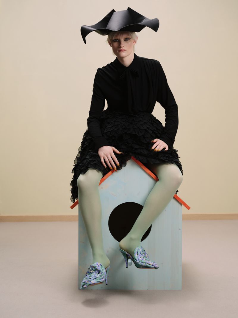 Stylish model in black hat, dress, and pastel tights seated on a whimsical painted dog house in a minimalist studio setting.