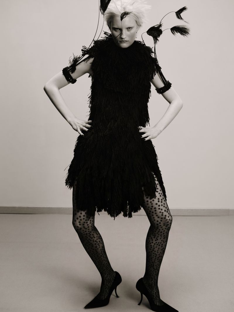 Woman in black feathered dress and heels strikes pose against neutral backdrop in fashion forward editorial shot with feathered details.