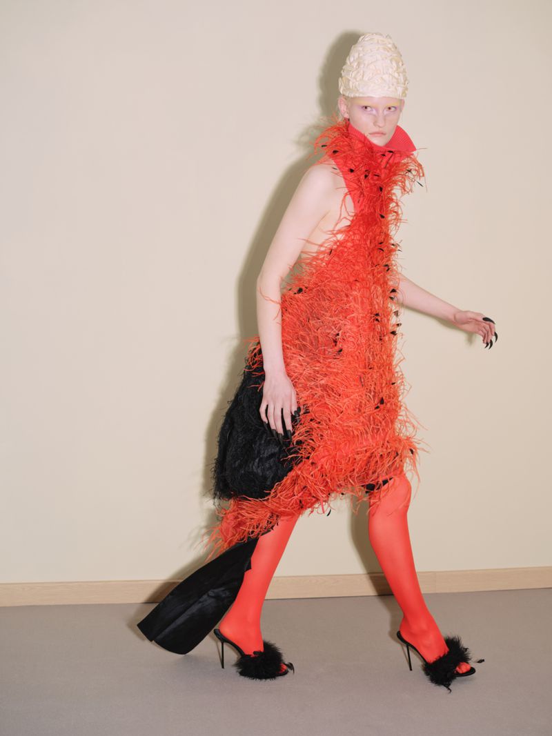 A slender fashion model walks in a sculptural red feathered dress, matching red tights and black heels against a neutral background.