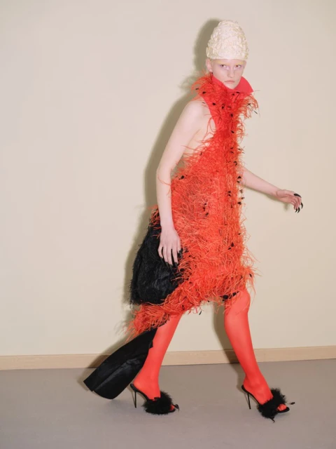 A slender fashion model walks in a sculptural red feathered dress, matching red tights and black heels against a neutral background.