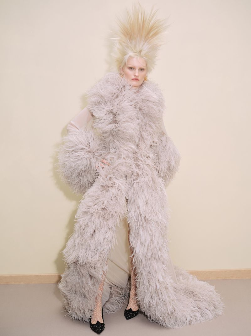 Fashion model with platinum blonde hair and feathery coat poses in studio, showcasing editorial haute couture fashion and dramatic styling.