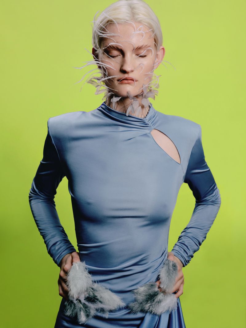 Fashion image of a platinum blonde model in a blue dress with feather accents against a bright green backdrop in a studio setting.