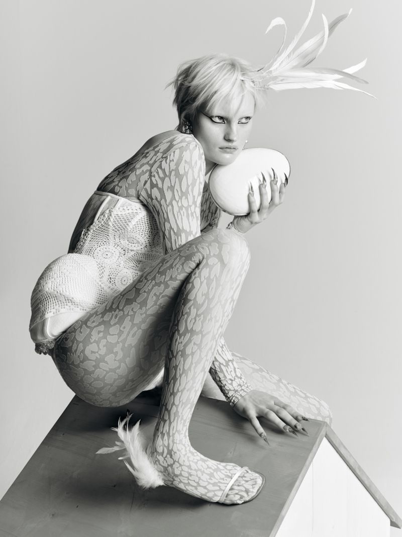 Conceptual fashion photo of a crouched woman in textured bodysuit holding a white egg on a birdhouse, against a white background.