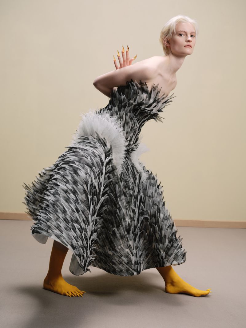 A blonde model with long gold-tipped fingernails wears a strapless black and white feathered dress with yellow socks, posing in a studio.