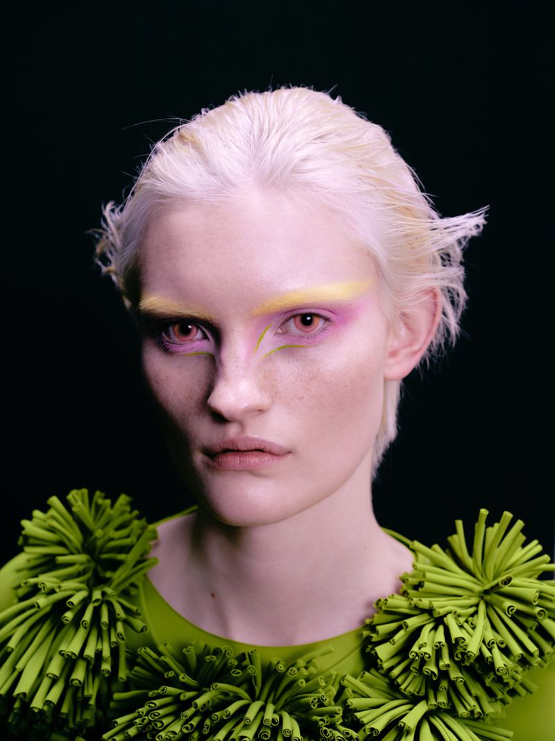 Close-up portrait of a light-skinned young adult woman with platinum hair, and creative makeup, wearing a green costume against a dark background.
