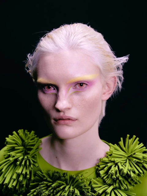 Close-up portrait of a light-skinned young adult woman with platinum hair, and creative makeup, wearing a green costume against a dark background.