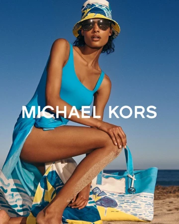 A woman poses on a beach modeling a turquoise bathing suit, sunglasses, and a patterned hat for Michael Kors, with a coordinated beach bag.