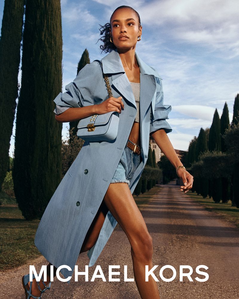 Elegant woman in a blue trench coat carrying a purse on a rural road lined with trees, a Michael Kors fashion ad.