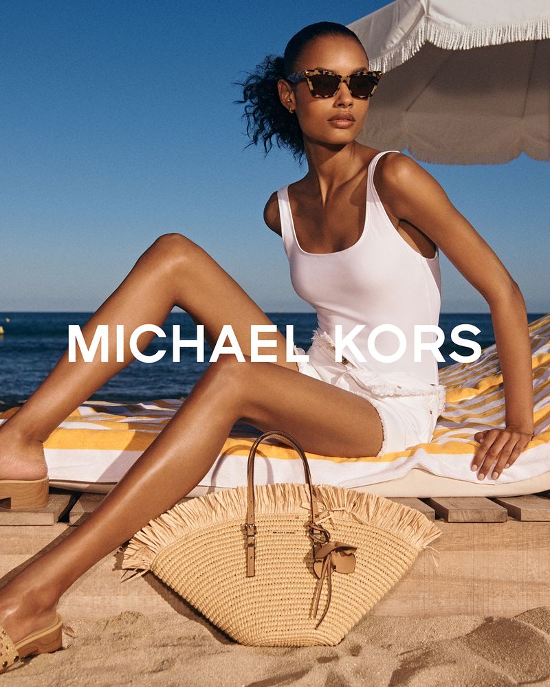 A woman poses on a beach next to a Michael Kors straw tote bag, wearing a white swimsuit top, white shorts, and tortoiseshell sunglasses.