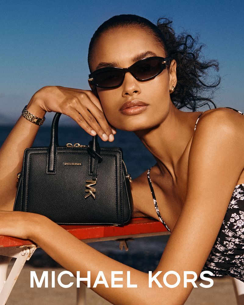 Elegant woman poses with a Michael Kors handbag and sunglasses against a clear blue sky backdrop, showcasing fashion and style.