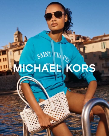 A woman models Michael Kors on a boat, wearing a turquoise hoodie, sunglasses, and carrying a white woven handbag with a Mediterranean view.