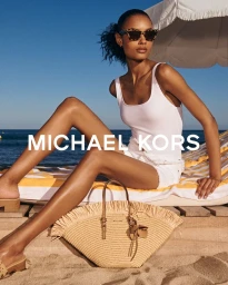 Model in white tank top and sunglasses seated on a sun lounger at the beach with a straw tote bag in the foreground.