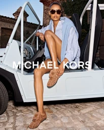 Model wearing oversized sunglasses, striped shirt, and brown shoes seated in a light blue utility vehicle outdoors