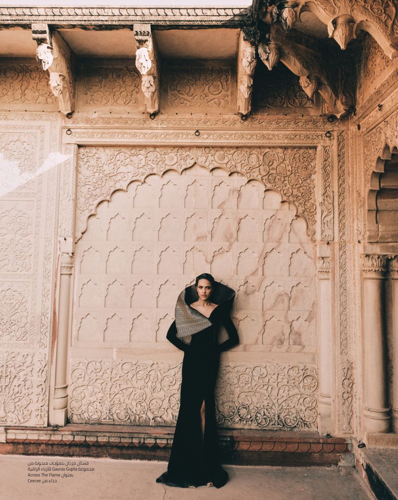 Fashion editorial shot of a woman in a black gown with an oversized collar, posing against an ornate wall in an architectural setting.
