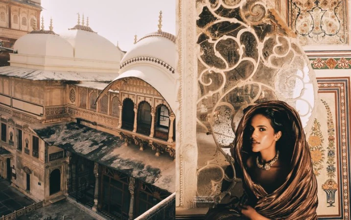 Woman in metallic fabric with an ornate necklace and historic architecture in the background. Architectural details are seen on the facade.