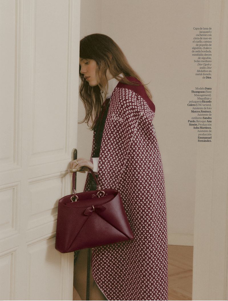 Brunette woman in patterned coat with burgundy purse, standing in doorway; fashion editorial shot with neutral indoor setting.