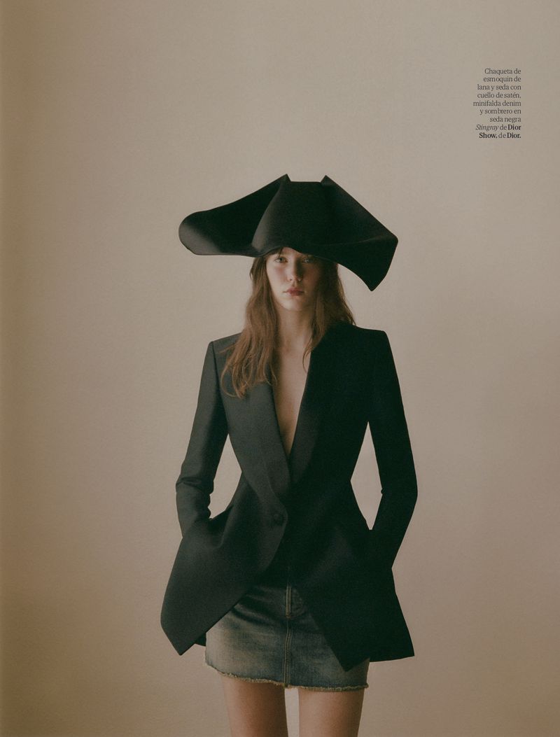 Fashion shot of a model wearing an oversized black hat, blazer and mini skirt against a neutral background, with magazine text visible.