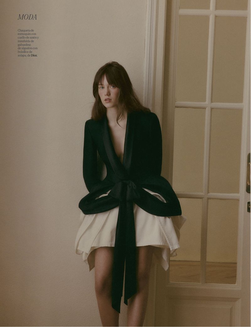 Adult female fashion model poses indoors wearing a dark green jacket with a large bow and a short white skirt next to a white door.