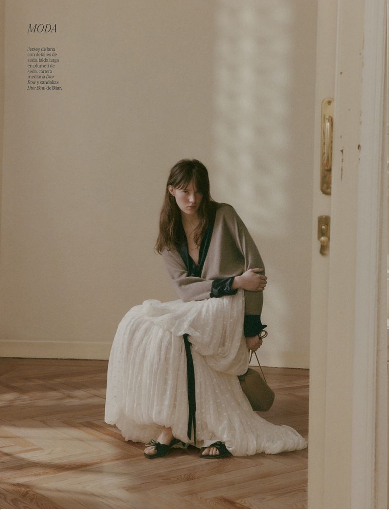 Fashion editorial shot of a woman in a beige cardigan and white pleated skirt sitting on a chair in a minimalist interior with warm lighting.