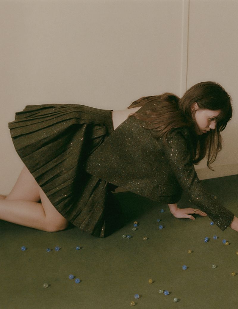 Woman in dark green skirt and jacket on green surface with scattered beads, laying down in an indoor setting with neutral background.