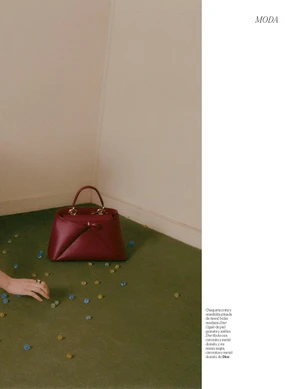 Burgundy leather handbag resting on a green carpet with scattered beads next to a hand in an editorial setting