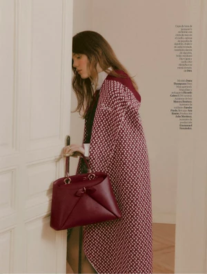 Model leaning against a white door holding a burgundy leather handbag wearing patterned coat