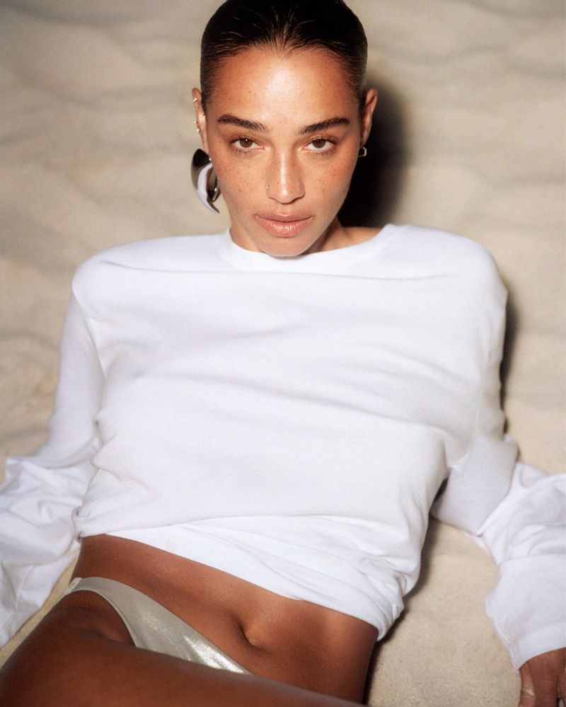 Portrait of a brunette woman in a white long-sleeve top with bare midriff, lying on a cream-colored sandy surface, indoor shot.
