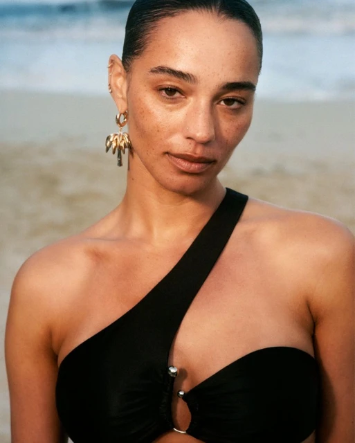Close-up of a tanned woman with dark hair, wearing a black swimsuit and palm tree earrings, posing on a tan sandy beach.